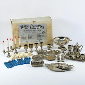 May include: A silver-colored miniature dining set with a variety of pieces, including plates, cups, a teapot, and serving dishes. The set includes a candelabra with red-tipped candles, silverware, and a box labeled "Terri's Treasures Silvercast Elegant Settings."