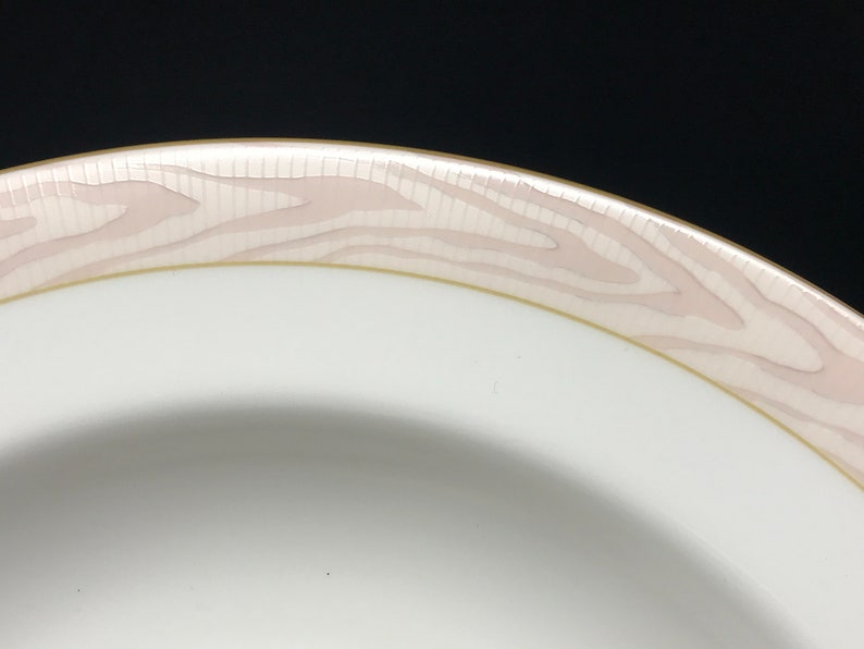 May include: Close-up of the rim of a white ceramic bowl with a pink and gold woodgrain design.