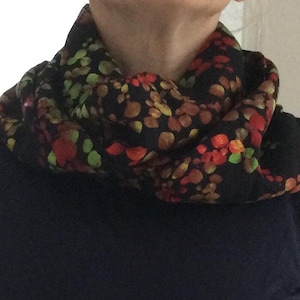 Könnte beinhalten: Ein schwarzer Loop-Schal mit einem bunten Blumenmuster. Der Schal hat rote, orangefarbene und grüne blattartige Formen auf dunklem Hintergrund. Der Schal ist um den Hals gewickelt.