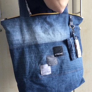 Könnte beinhalten: Eine blaue Tragetasche aus Jeansstoff mit gelbem Reißverschluss, zwei Schulterriemen und drei quadratischen Stoffaufnähern mit den Texten STRONG, COSY und just be. Eine kleine aufgesetzte Jeanstasche ist mit einem dekorativen Stoffanhänger versehen.