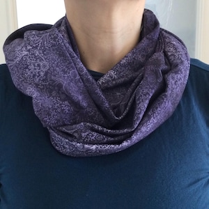 Könnte beinhalten: Ein violetter Rundschal mit detailliertem Paisley-Muster. Der Schal ist um den Hals gewickelt und aus einem weichen Stoff gefertigt. Der Hintergrund ist ein dunkelblaues Oberteil.