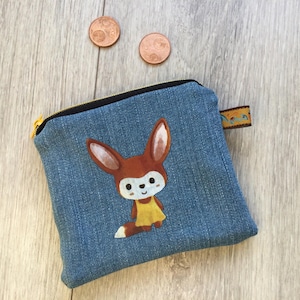 Könnte beinhalten: Eine blaue Jeans-Reißverschlusstasche mit einer Cartoon-Fuchs-Illustration. Der Fuchs hat braune Ohren und ein gelbes Kleid. Die Tasche hat einen schwarzen Reißverschluss und eine kleine Stoffschlaufe. Zwei Kupfermünzen sind sichtbar.