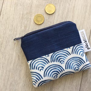 Könnte beinhalten: Ein kleines, marineblaues Münzportemonnaie mit Reißverschluss. Die untere Hälfte hat einen weißen Stoff mit blauem Wellenmuster. Zwei Goldmünzen liegen in der Nähe des Portemonnaies. Ein Etikett lautet "glück!ich".