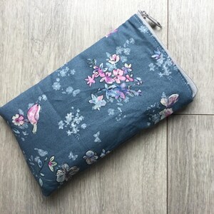 Könnte beinhalten: Eine rechteckige, denimfarbene Reißverschlusstasche mit floralem Muster. Das Muster zeigt rosa, lila und weiße Blumen sowie kleine Schmetterlinge und einen Vogel. Die Tasche liegt auf einer hellen Holzmaserung.
