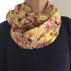 Könnte beinhalten: Ein gesteppter Loop-Schal mit einem botanischen Muster. Der Schal hat ein sich wiederholendes Design aus Blättern in Gelb-, Rosa- und Orangetönen auf einem hellgelben Hintergrund. Der Schal ist um den Hals gewickelt, und die Person trägt ein dunkelblaues Langarmshirt.