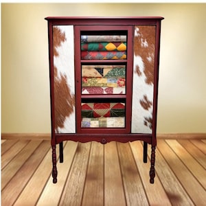May include: A red wooden cabinet with a glass door and two solid doors. The cabinet has a cowhide pattern on the doors and shelves filled with colorful patchwork quilts.