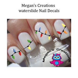 May include: White nails with Christmas lights nail decals. The lights are red, green, blue, yellow, and orange. The decals are on a white background. The image also includes a small owl logo with the text "Megan's Creations".