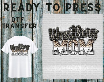 May include: White t-shirt with a "Wrestling Mom" design. The design features the word "Wrestling" in a script font with a leopard print pattern, and "MOM" in a block font. The image also includes the text "READY TO PRESS" and "DTF TRANSFER".