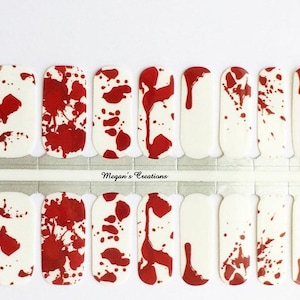 May include: A set of nail polish strips featuring a white base with a red splatter pattern resembling blood drops. The strips are arranged in two rows on a backing sheet with the text Megan's Creations printed in the center of the strip divider.