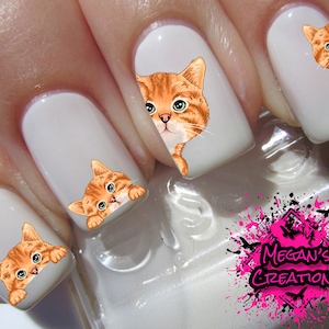 May include: Close-up of a hand with several fingernails decorated with orange cat nail art stickers. The stickers feature orange tabby cats peeking out from behind the nail.
