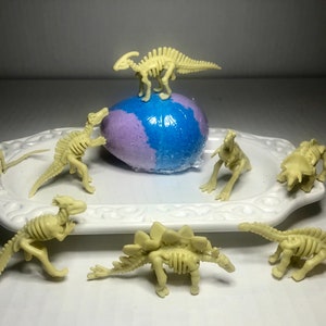 May include: A collection of seven small, beige plastic dinosaur skeletons on a white plate. One dinosaur is standing on top of a purple and blue bath bomb.