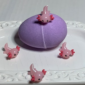 May include: Four pink and white axolotl figurines with red gills. The axolotls are sitting on a white plate with a purple bath bomb in the center.
