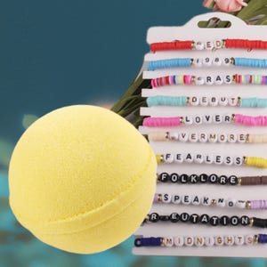 May include: A bright yellow bath bomb sits next to a display of colorful beaded bracelets. The bracelets have white letter beads spelling out words like "RED", "LOVER", and "MIDNIGHTS". The background is a teal color.