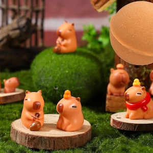 May include: A collection of miniature figurines of capybaras in various poses, some with small yellow hats, on a green moss surface. The figurines are made of brown and orange plastic and are about 2 inches tall.
