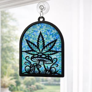 Op de afbeelding: Zwart omrande raamhanger met een cannabisblad- en paddestoelontwerp. Het kunstwerk heeft een zwart silhouet tegen een blauw en groen patroon. De raamkunst wordt opgehangen door een zuignap.
