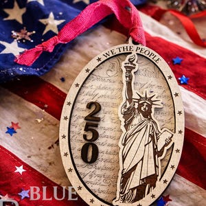May include: Wooden oval ornament featuring the Statue of Liberty, the text "WE THE PEOPLE", and the number "250". The ornament is set against a backdrop of an American flag and a pink ribbon.