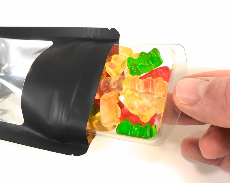 Peut inclure: Un sachet refermable noir avec une fen&ecirc;tre transparente, partiellement ouvert, r&eacute;v&eacute;lant un r&eacute;cipient en plastique transparent rempli d'oursons g&eacute;lifi&eacute;s color&eacute;s.