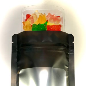 Peut inclure: Un sachet refermable noir avec une fen&ecirc;tre transparente, rempli de bonbons g&eacute;lifi&eacute;s color&eacute;s en forme d'ours. Le sachet est pos&eacute; sur une surface blanche.