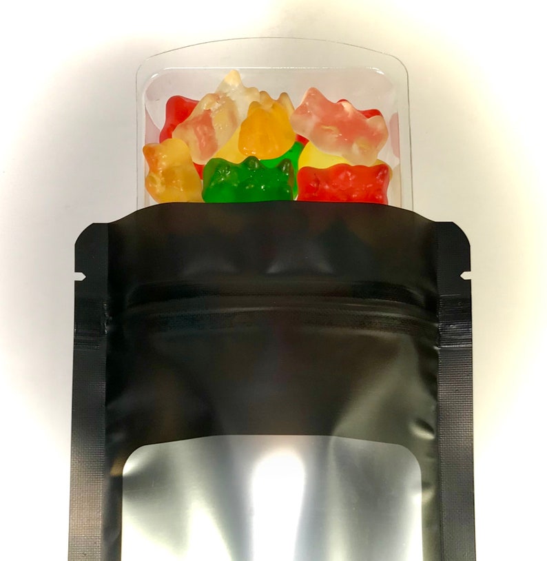 Peut inclure: Un sachet refermable noir avec une fen&ecirc;tre transparente, rempli de bonbons g&eacute;lifi&eacute;s color&eacute;s en forme d'ours. Le sachet est pos&eacute; sur une surface blanche.