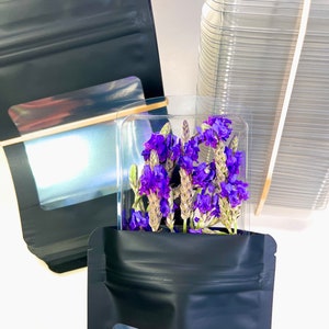 Peut inclure: Deux sachets debout noirs avec des fen&ecirc;tres transparentes, l'un contenant un bouquet de fleurs violettes. Les sachets sont &agrave; c&ocirc;t&eacute; d'une pile de contenants en plastique transparents.