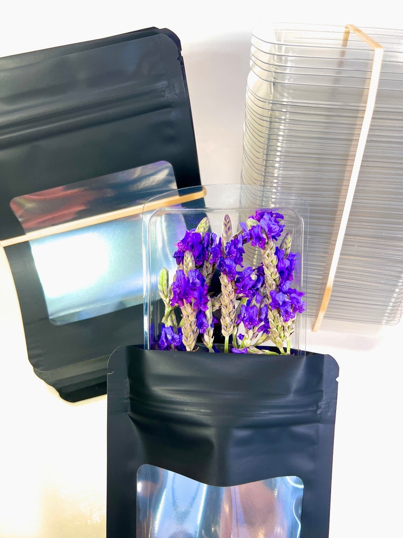 Peut inclure: Deux sachets debout noirs avec des fen&ecirc;tres transparentes, l'un contenant un bouquet de fleurs violettes. Les sachets sont &agrave; c&ocirc;t&eacute; d'une pile de contenants en plastique transparents.
