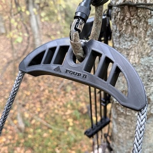 May include: A gray bow holder with the text "FOUR B" is attached to a tree. The bow holder has a series of rectangular cutouts and is connected to a rope. The background is blurred, showing a forest setting.