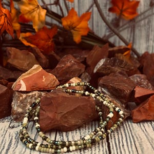 May include: A beaded necklace with a mix of green, white, and black beads. The necklace is displayed on a bed of red rocks, with orange and yellow fall leaves in the background. The beads are small and round.
