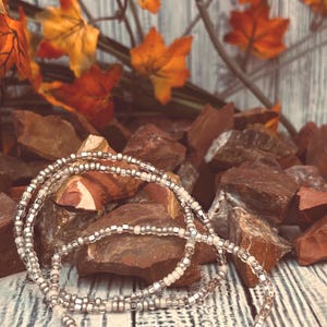 May include: Three strands of beaded jewelry with silver and white beads. The jewelry is displayed on a wooden surface with brown rocks and orange autumn leaves in the background. The beads are small and round.