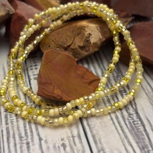 May include: A collection of beaded waist beads in shades of yellow, cream, and silver. The beads are strung together, creating a layered effect. The beads are displayed on a wooden surface with a rock background.