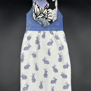 May include: A white kitchen towel with a blue and black floral print hanging loop. The towel has a pattern of gray rabbits on a white background.
