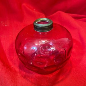 May include: A spherical, red glass bottle with a textured design and a dark green metal rim. The bottle rests on a red fabric. This could be a decorative item or a container for liquids.