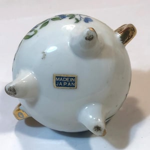 以下が含まれることがあります： 3つの金色の足と取っ手が付いた、小さな白いセラミックボウル。ボウルは緑と青の花柄で装飾されています。底の長方形のステッカーには「MADE IN JAPAN」と書かれています。