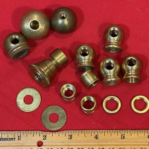 Puede incluir: Una colección de piezas de lámpara de latón vintage, que incluye remates esféricos, enchufes roscados, arandelas y piezas de conexión. Los componentes están dispuestos sobre una superficie roja, con una regla para la escala.