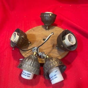 May include: A circular wooden ceiling light fixture base featuring four bronze-toned metal lamp sockets with intricate leaf-like patterns. Each socket includes white ceramic interiors with text reading 125V, 2A, 300W and USE 60 W MAX.