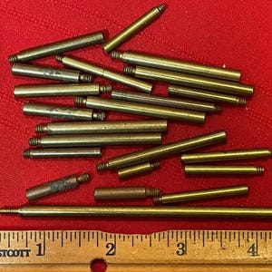 May include: A selection of brass-coloured metal rods of different lengths, some with threaded ends, arranged on a red surface. A wooden ruler with inch markings is at the bottom.