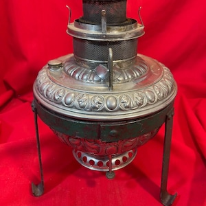 May include: A silver-toned metal oil lamp burner featuring an ornate embossed floral pattern on the rounded body. The top section includes a cylindrical mesh chimney component. It is supported by a dark green metal tripod stand with curved feet.