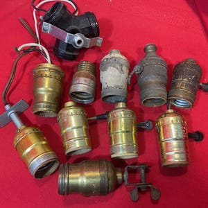 May include: A collection of vintage brass and black lamp sockets and components, including a three-way socket, various bulb holders, and a clamp. The items are arranged on a red surface, showcasing their aged appearance and diverse designs. Ideal for restoration or repurposing projects.