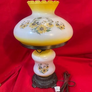 May include: A hurricane-style table lamp featuring a white and yellow frosted glass shade and base, both decorated with a sunflower and foliage pattern. The lamp has a dark brown, ornate metal base and a brown electrical cord with a white plug.