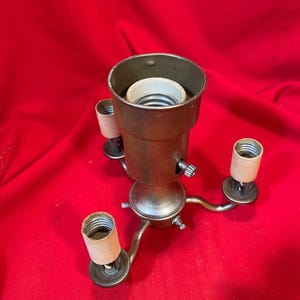 Könnte beinhalten: Ein Vintage-Lampensockel mit Bronze-Finish. Der Lampensockel hat eine zentrale Glühbirnenfassung und drei kleinere Fassungen, die sich nach außen erstrecken. Die Basis wird von drei gebogenen Armen getragen. Der Hintergrund ist einfarbig rot.