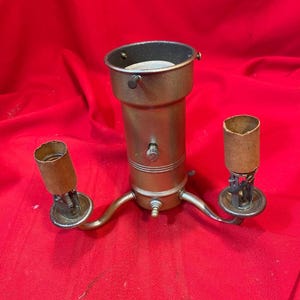 Könnte beinhalten: Vintage Lampenfassung in Messingoptik mit zwei seitlichen Fassungen und einer zentralen Fassung. Die Fassung hat einen zylindrischen Körper mit einem Schalter und einem dreibeinigen Sockel. Die Fassungen sind zylindrisch und aus einem ähnlichen Messingfarbenen Metall.