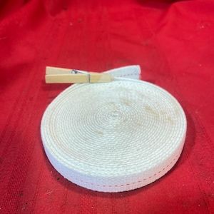 May include: A coiled roll of off-white woven cotton webbing tape featuring a thin red stitched line along the edge. The roll is secured with a small wooden clothespin that clips the end of the tape to the main body of the coil.