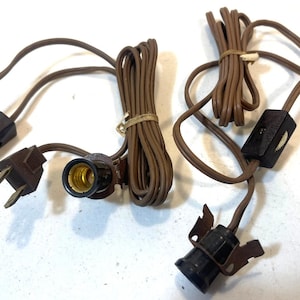May include: Several brown lamp cords with plugs, switches, and bulb sockets are arranged on a white surface. The cords are bundled and coiled, with the switches and plugs in a dark brown color. The bulb sockets have a gold-colored interior.