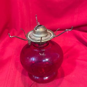 Könnte beinhalten: Ein antiker Öllampenfuß aus durchscheinendem rubinrotem Glas mit bauchigem Körper und ausgestelltem Standfuß. Oben befindet sich ein runder Metallbrenner mit vier gebogenen Drahtarmen, die zur Halterung eines Glasschirms dienen.