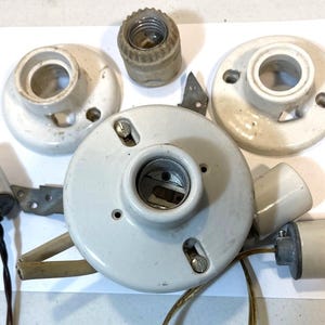 Może przedstawiać: Kolekcja zabytkowych elementów elektrycznych, w tym białe ceramiczne oprawki żarówek, gniazdo żarówki i oprawa wielogniazdowa. Widoczne są również przewody i metalowe wsporniki, co sugeruje naprawę lub renowację elektryczną.