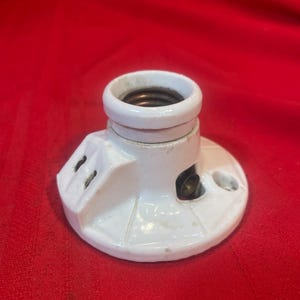 Puede incluir: Un portalámparas de cerámica blanca con base circular y parte superior cilíndrica. El portalámparas tiene dos pequeños orificios y un contacto metálico en el interior. El fondo es de color rojo liso.