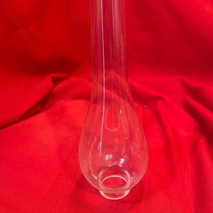 May include: A clear glass lamp chimney with a rounded, bulbous shape and a flared top. The chimney is transparent, allowing light to pass through. It sits on a red background.
