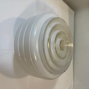 Könnte beinhalten: Eine weiße und transparente Glasleuchte mit einem Spiraldesign. Die Leuchte hat eine abgerundete Form mit vertikalen Rippen an den Seiten und einem kreisförmigen Muster in der Mitte. Die Leuchte ist wahrscheinlich für eine Decke oder Wand.