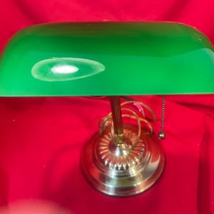 May include: A classic green banker's lamp with a brass base and a pull chain. The lamp features a curved, green glass shade and a decorative base. The lamp is designed for desk or table use and provides focused lighting.