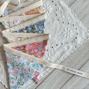 May include: A colorful fabric bunting with a floral pattern. The bunting is made of five triangular flags sewn together with a cream-colored fabric ribbon. The bunting has a tag that says "elliebug makes".