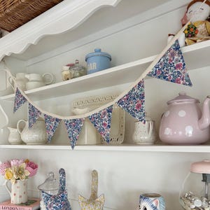 May include: A floral patterned bunting with blue, pink, and white flowers hangs above a shelf filled with decorative items. The shelf displays a pink teapot, a blue sugar container, and cat-shaped fabric ornaments.